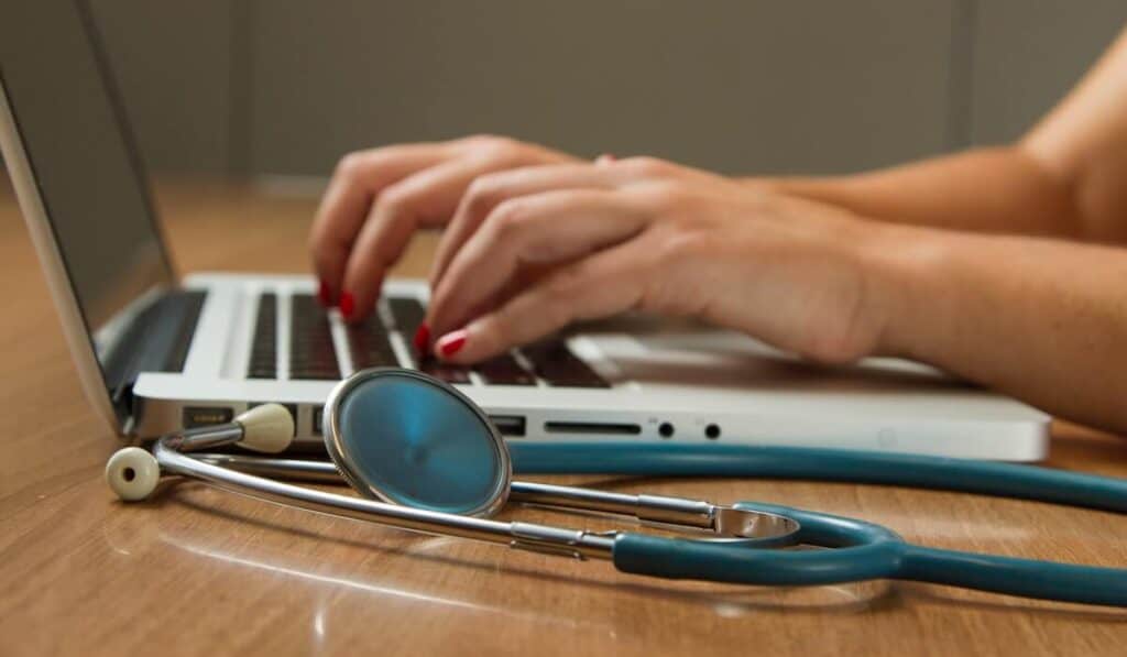 Medical professional typing on a laptop with a stethoscope on the table next to them